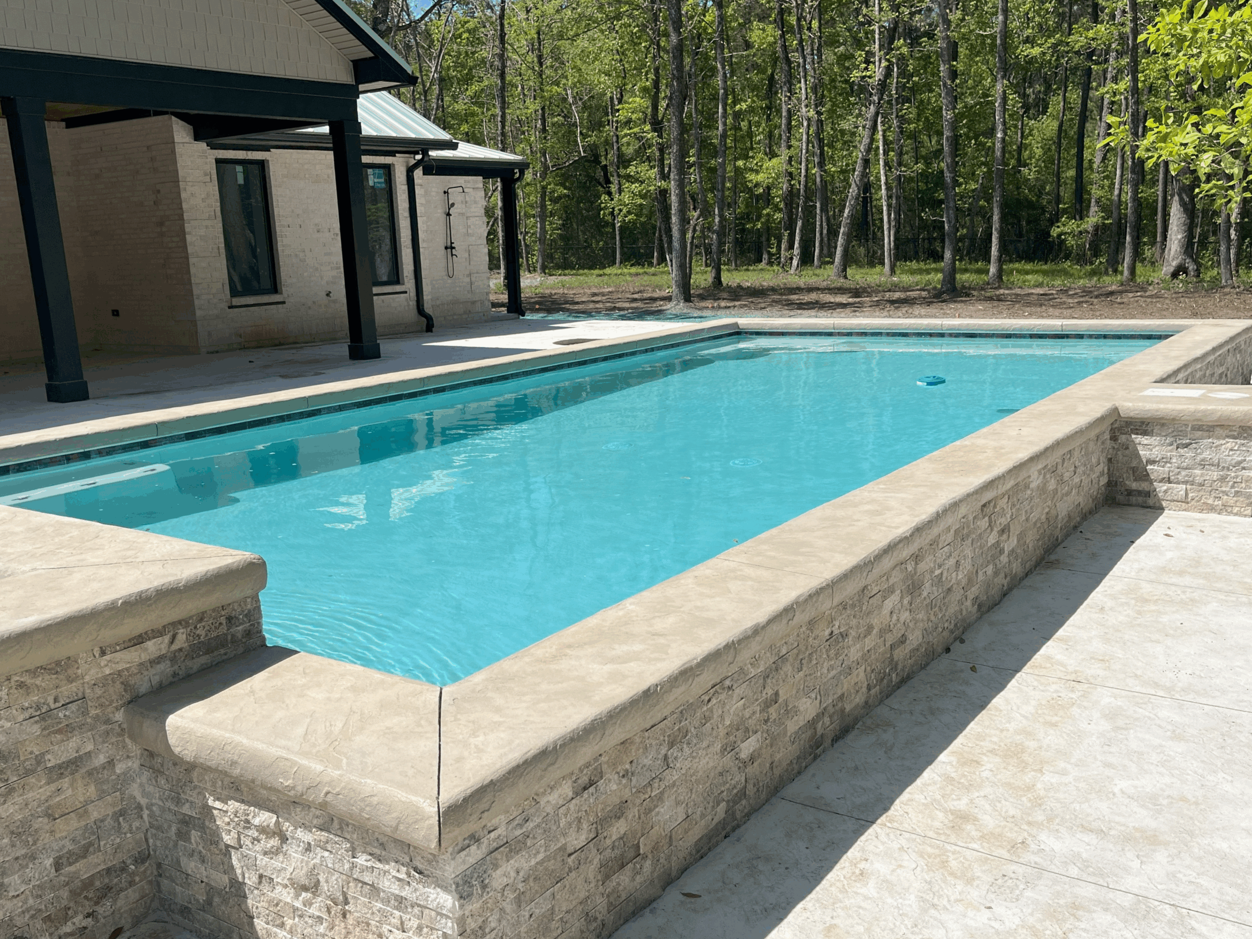 A gunite pool with low sides with stone exteriors. The pool is surrounded by a concrete patio and there are trees in the background.